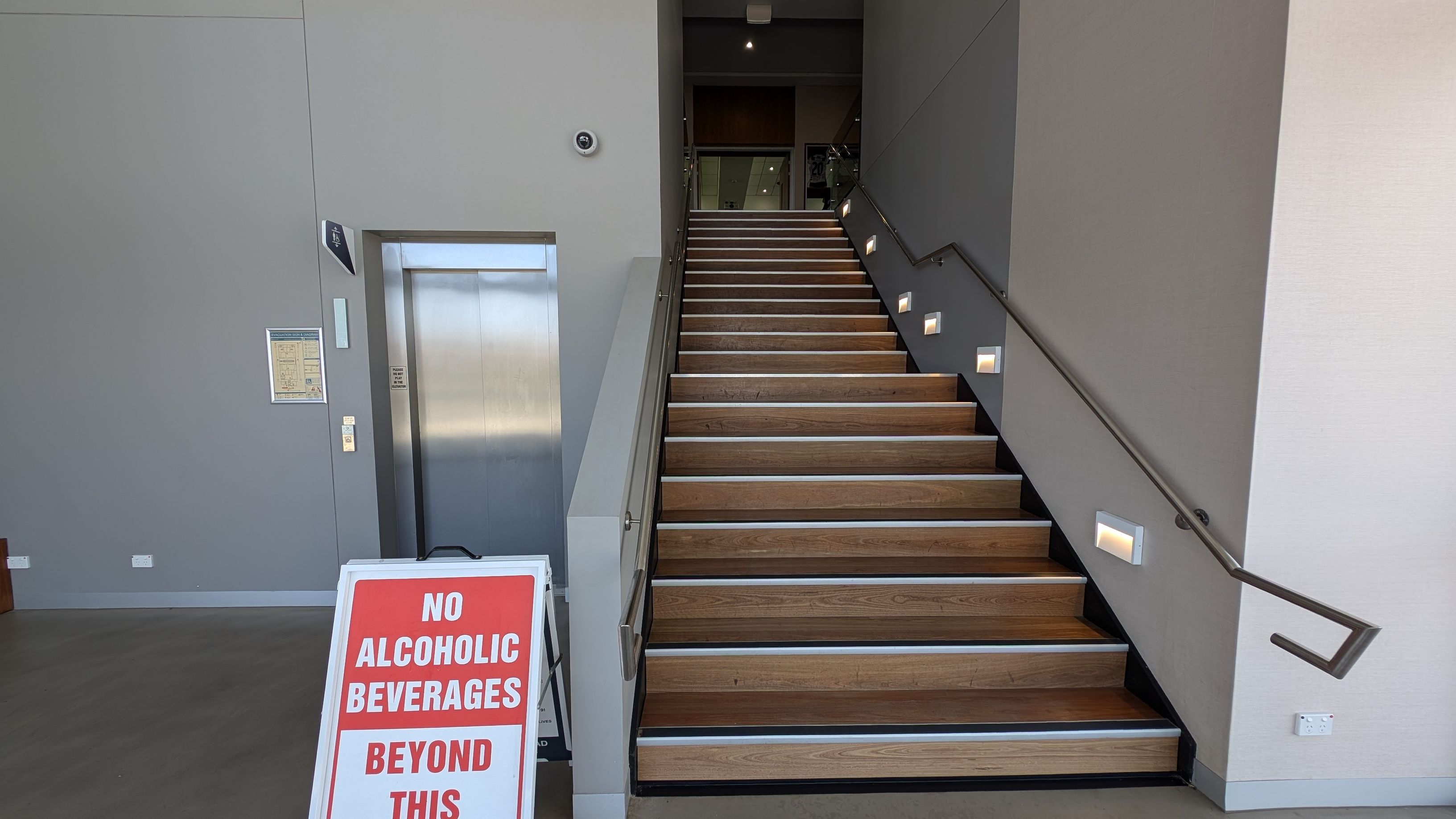 A modern interior with a grey floor and walls. On the left, there's a metallic lift with control buttons. A staircase with wooden steps and a black handrail is in the centre, with small lights on each step. In front of the stairs, a sign reads "No Alcoholic Beverages Beyond This Point." On the right, large glass doors and windows reveal a view of greenery and a clear sky outside. The ceiling is not visible.