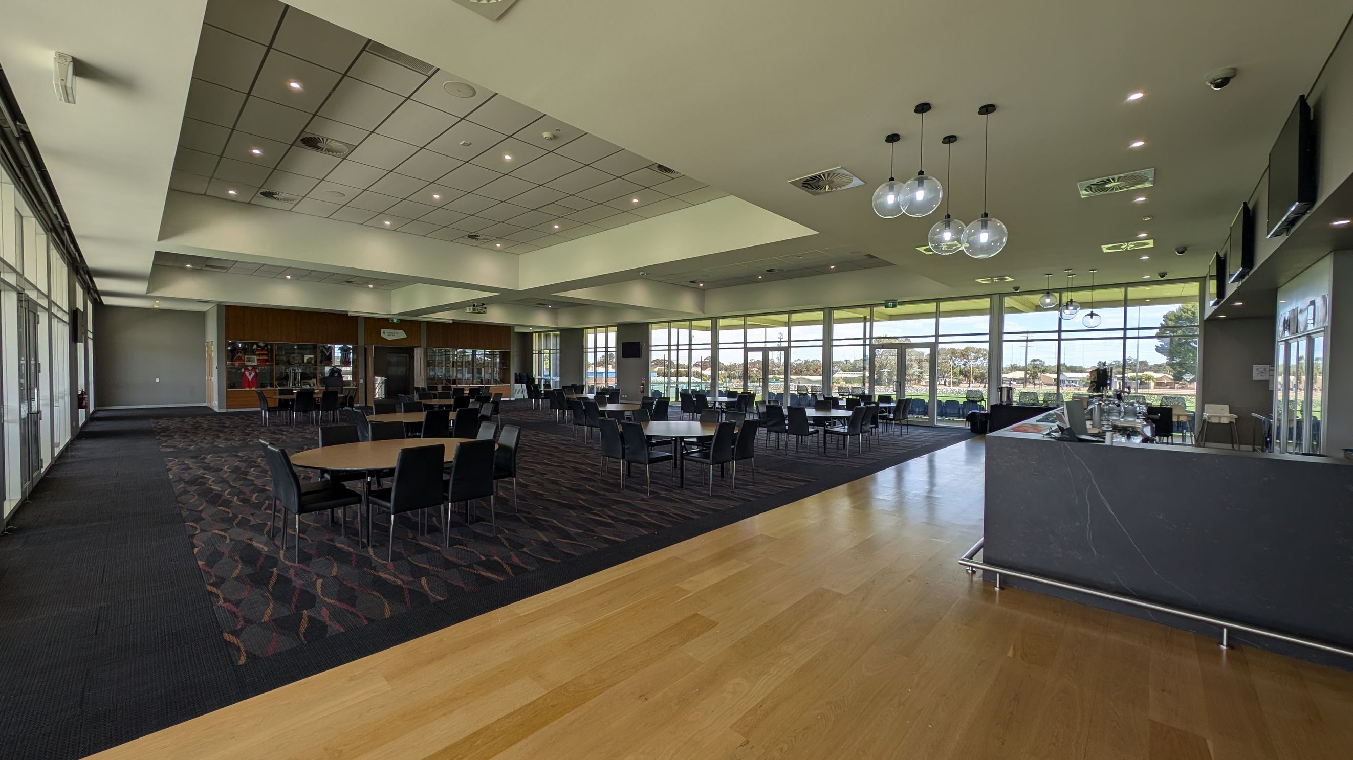 A spacious, modern dining area with a mix of carpet and wooden flooring. On the right, there's a sleek bar counter with hanging spherical lights above. The left side features rows of tables and chairs, with a wall of windows in the background allowing natural light to flood in. The ceiling is fitted with recessed lighting and air vents. Large glass doors and windows provide a view of the outdoors.
