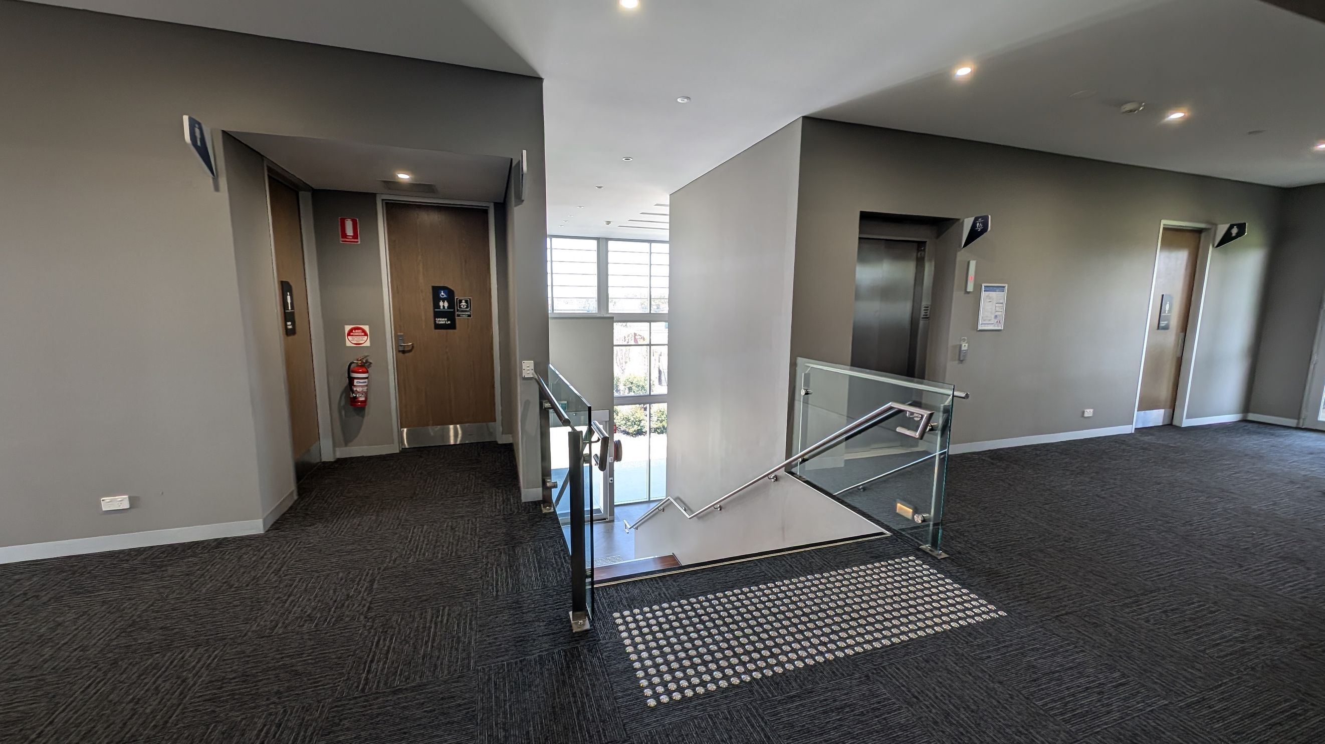 A modern indoor space with two lifts on either side of a central staircase. The left lift is next to a wooden door with a fire extinguisher mounted on the wall. The right lift is adjacent to a notice board. The staircase has glass railings and leads downwards, with a large window in the background allowing natural light. The floor is carpeted in dark grey, and the ceiling is fitted with recessed lights.