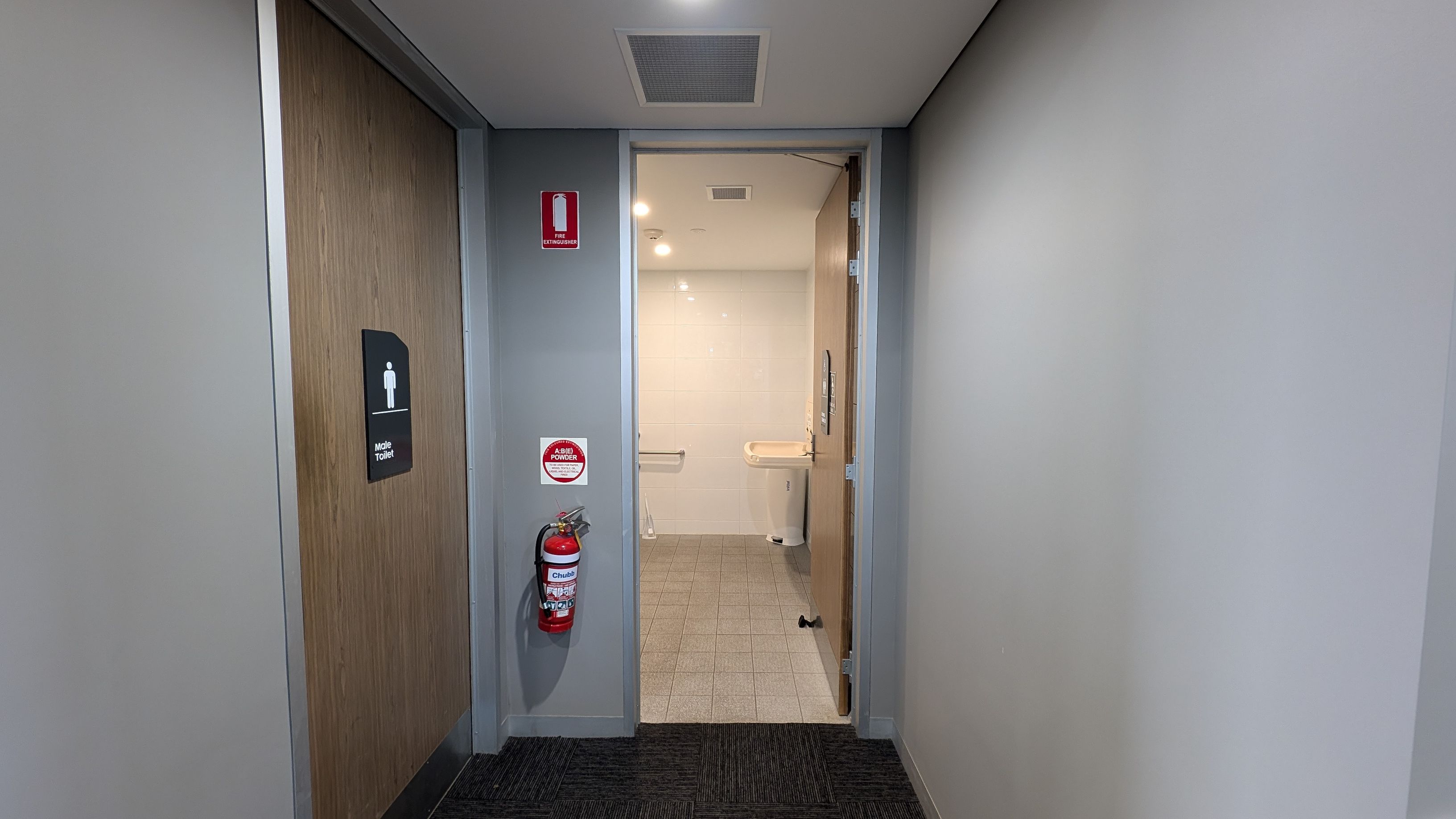 A hallway with grey walls and a dark carpeted floor. On the left, a closed wooden door with a sign indicating "Male Toilet." A fire extinguisher is mounted on the wall next to the door. The door on the right is open, revealing a bathroom with light-coloured tiles and a sink. The ceiling is white with a vent and recessed lighting.