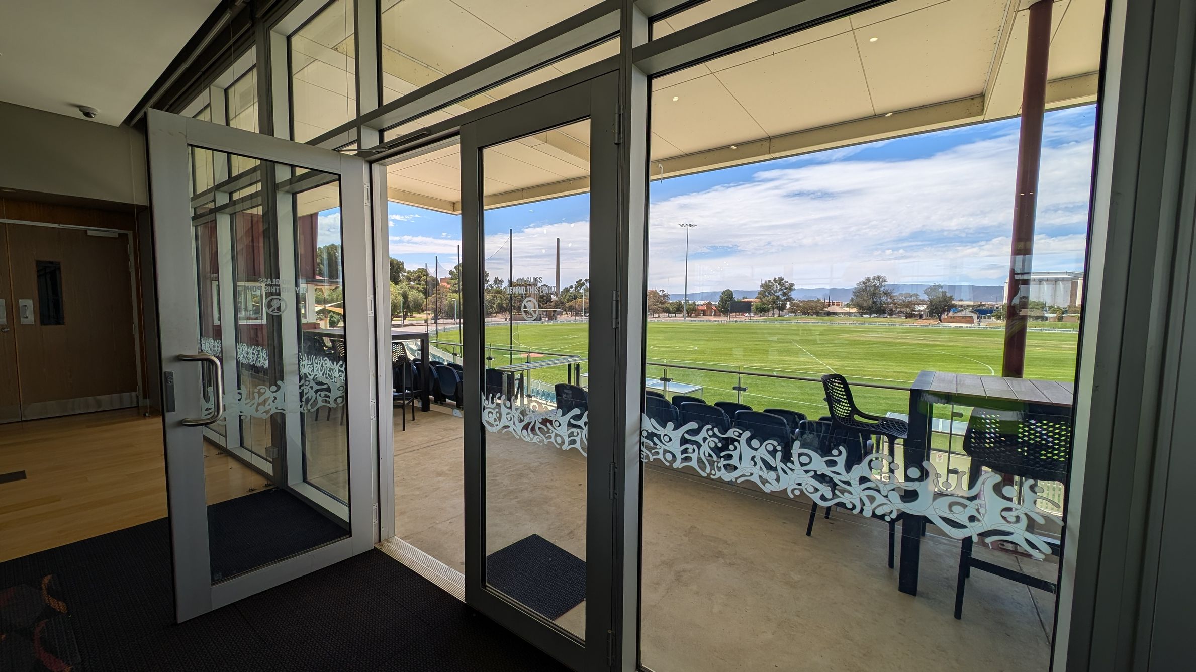 Glass doors open to a balcony overlooking a green sports field. The doors have decorative frosted patterns. On the balcony, there are several black chairs and a table. Beyond the field, trees and buildings are visible. The ceiling is light-coloured, and the floor inside is carpeted, transitioning to a concrete surface outside.