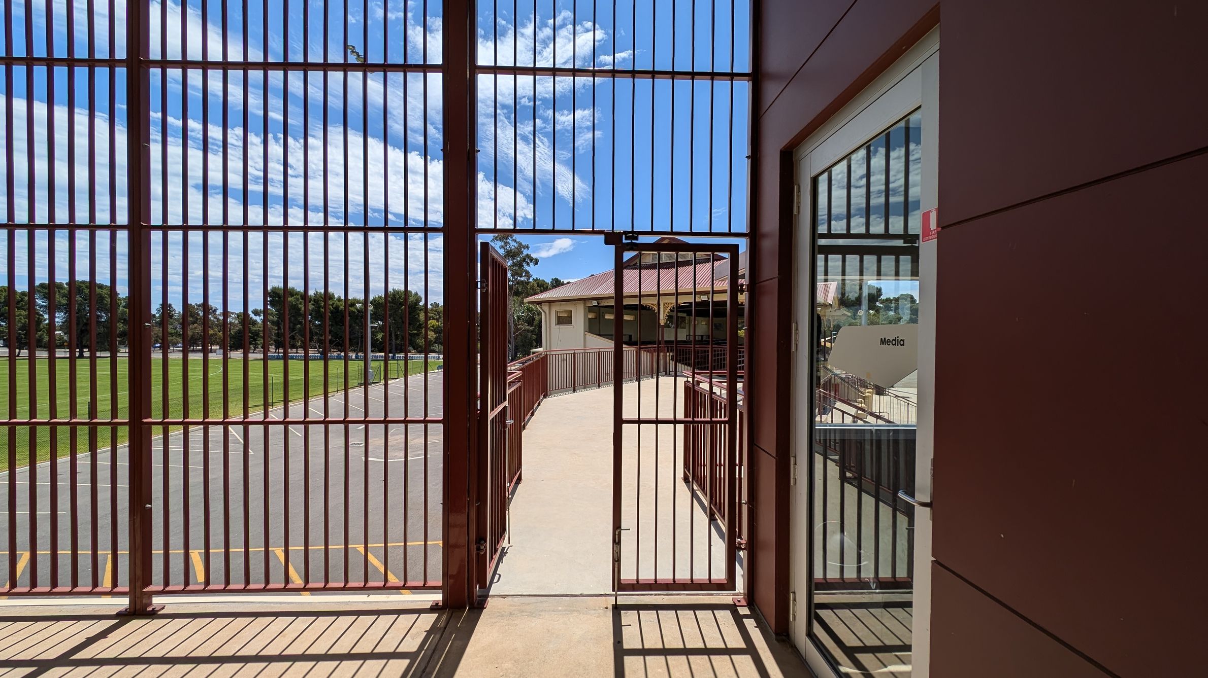 A view through a metal gate with vertical bars, leading to a concrete pathway. On the right, there's a glass door with a "Media" sign. Beyond the gate, a building with a red roof is visible. To the left, a grassy field and a paved area with yellow markings can be seen. Trees line the background. Shadows of the bars are cast on the ground.