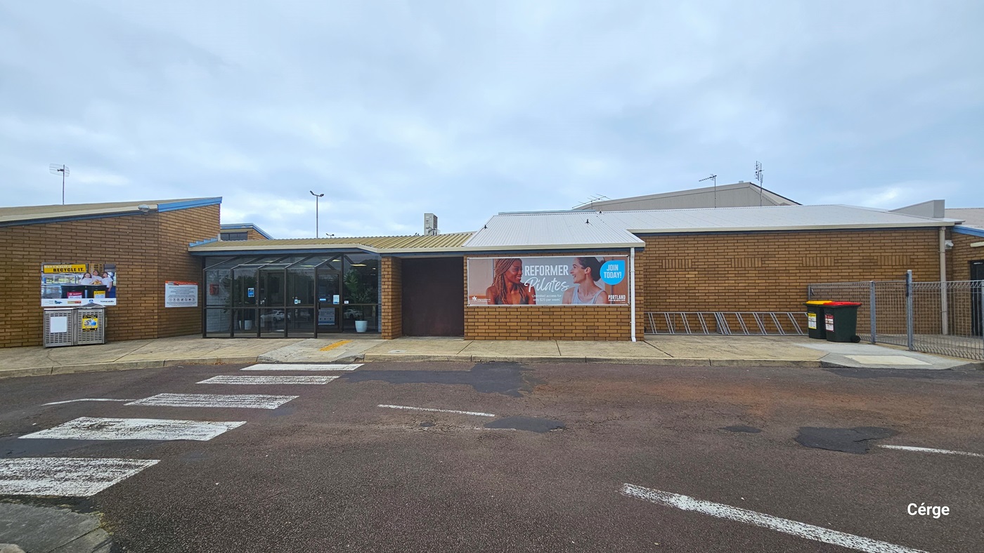 Accessible car parking spaces at Portland Leisure and Aquatic Centre. The regular parking spaces are marked by white lines, while the accessible car parking spaces are marked by yellow lines and are close to the facility, which is enclosed by a grey fence. The facility is a combination of brown brick walls, glass windows, and a white corrugated roof. Overhead, power lines stretch across the overcast sky.