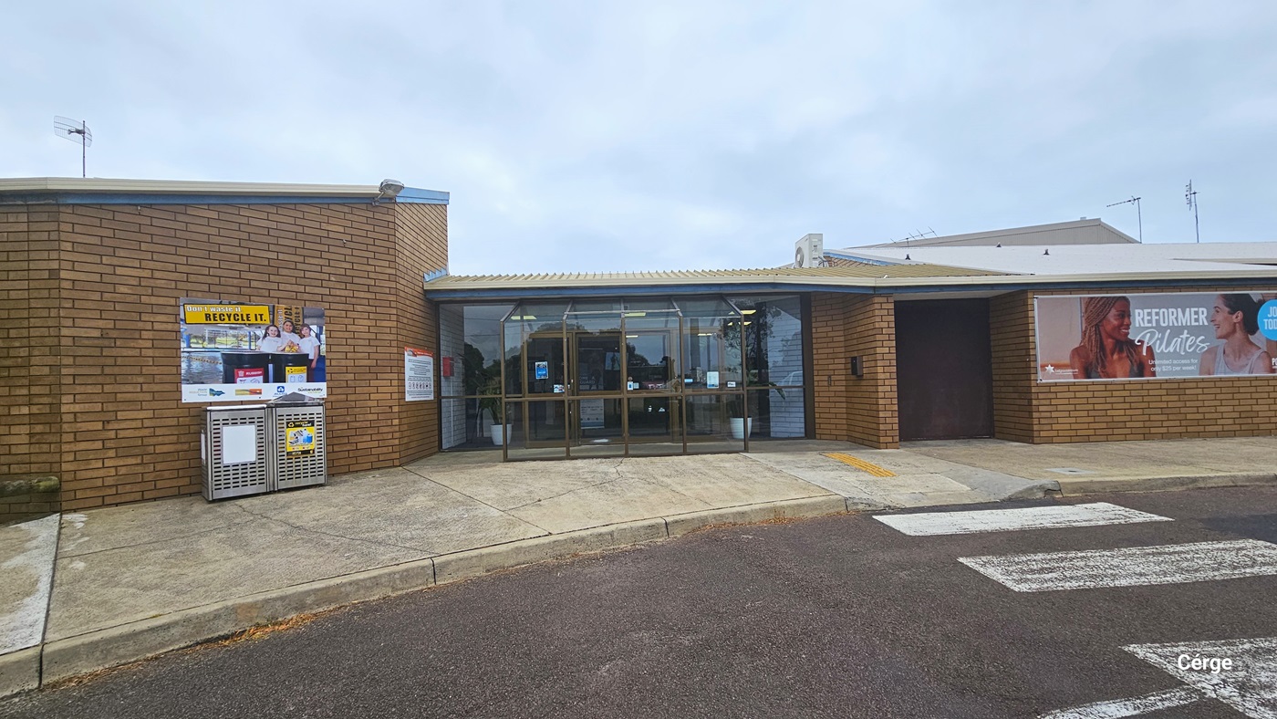 Concrete concourse of the low-rise, brown brick wall building with cream and white corrugated roof and a glass entrance with chocolate metal frames. On the left wall is a white sign, and further to the left are two stainless steel perforated garbage bins in front of the wall with a RECYCLE IT poster. On the glass doors are small infographics. Seen through the glass are white brick walls and white potted plants.