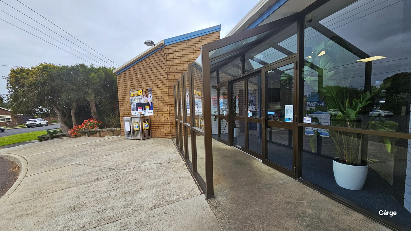 A closer look at the glass entrance doors with chocolate steel frames. Inside are two white potted plants that have banana-like leaves, a grey floor, and a white brick wall. Mounted on top of the left brown brick wall outside, right above the RECYCLE IT poster and perforated bins, is an outdoor lighting fixture. The facility has a garden with red flowers and tall trees with wide canopy cover.