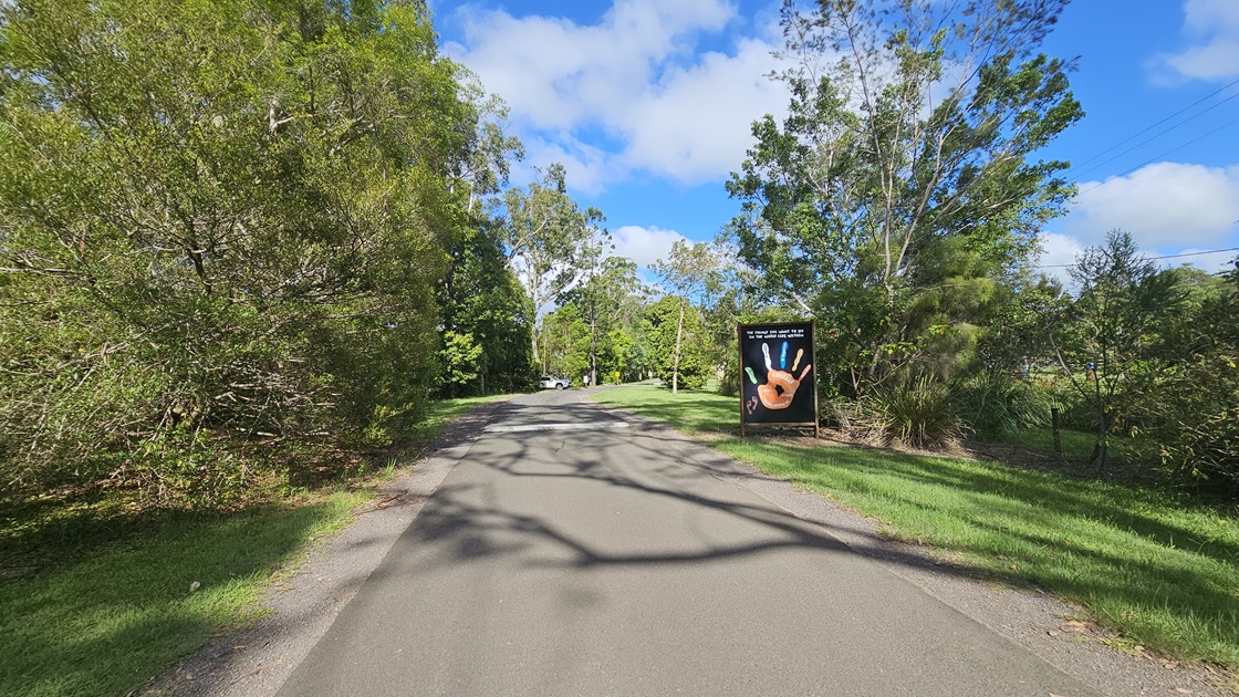 A straight bitumen road. Alongside the road, there is a large sign situated on the grassy area to the right. The road is flanked on both sides by various trees with robust branches and grass.