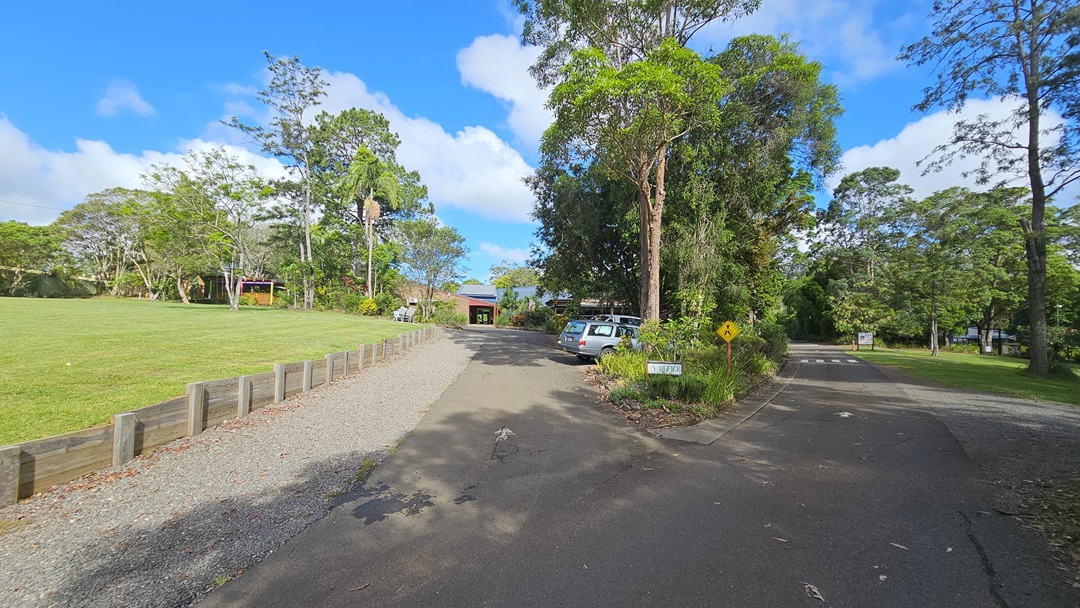 The road splits: To the left, it leads to a car park and a building with a brown brick facade, alongside a field of manicured grass bordered by a low concrete fence. The right-hand road leads to an open area surrounded by trees and grass. In the middle aisle, there is a sign, set against a backdrop of tall trees and grass.