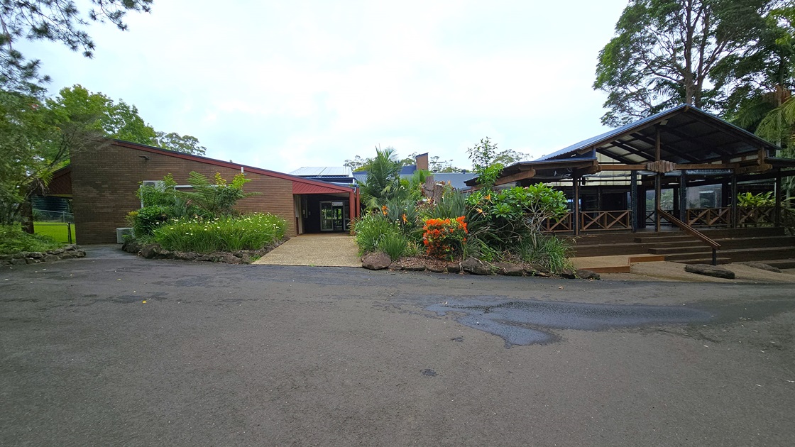 To the left, there's a low-rise building with a brown concrete facade and pitched roofing. It is surrounded by a landscaped garden with colourful flowering plants. On the right is another building with a pitched corrugated roof, accessible via concrete steps with railings. Wooden railings enclose this area. Tall trees of various types surround the vicinity.
