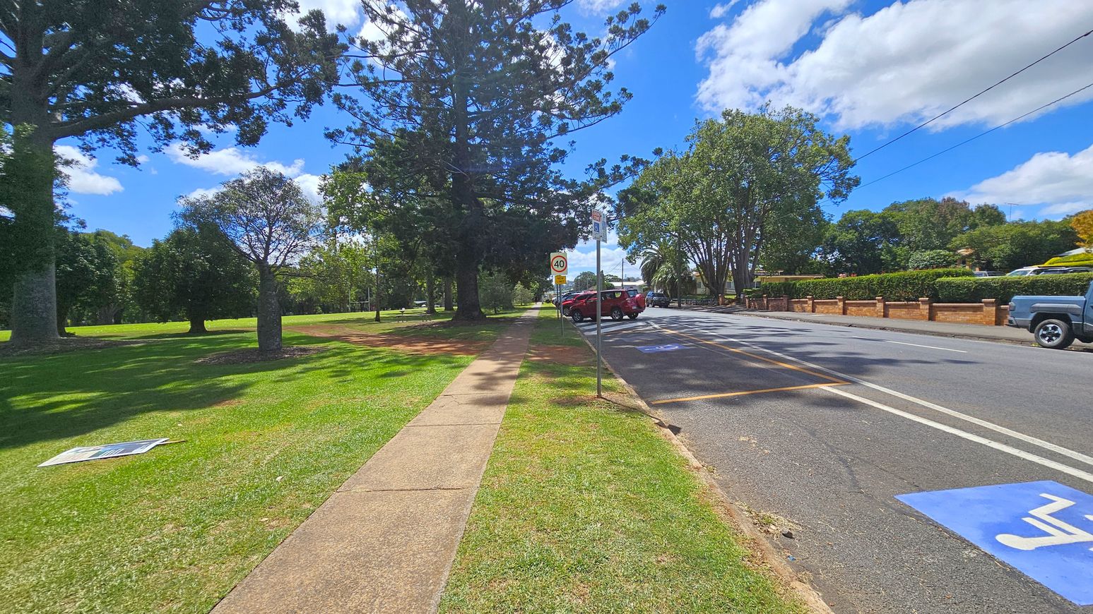 A paved footpath runs along the left side, bordered by a grassy area with several large trees providing shade. On the right, a road with marked parking spaces has a few parked cars. A speed limit sign indicates 40 km/h. A brick wall with hedges lines the far side of the road. The sky is partly cloudy, and the scene is well-lit, suggesting daytime.