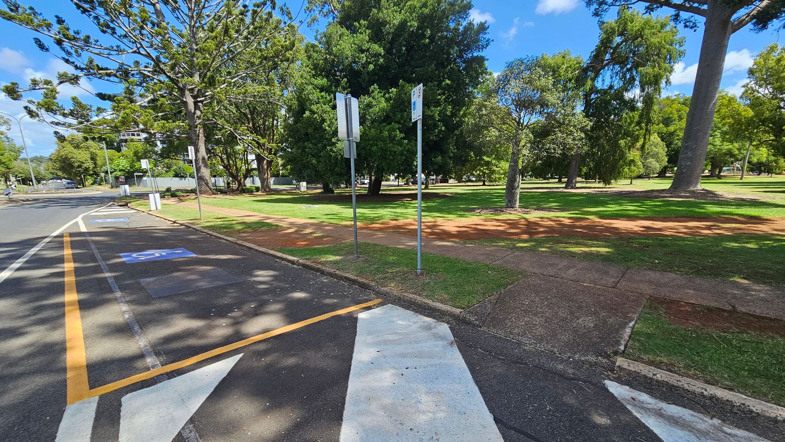 A paved road with marked parking spaces, including a disabled parking spot, is on the left. Two signs are visible near the kerb. On the right, a grassy park area with several large trees and a red dirt path is visible. The background features more trees and a building partially obscured by foliage. Shadows from the trees are cast on the road and grass.