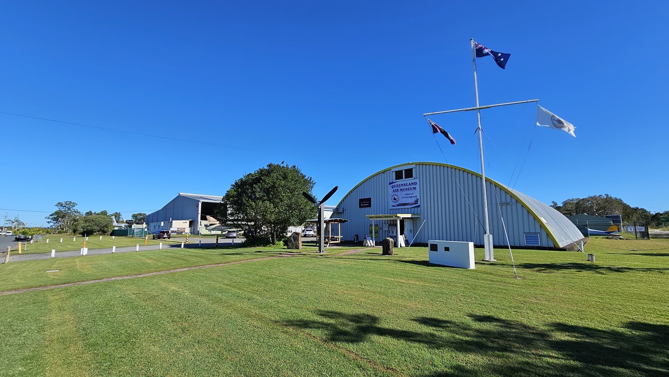 A substantial grey industrial structure is positioned behind a well-kept grass field. Near the centre and closer to the front, a smaller building with a domed metal roof displays a sign and a mounted propeller. An Australian flag, among other flags, flies on a pole to the right of this building. A neatly manicured grass area stretches in the foreground, with clear paths leading to the buildings.