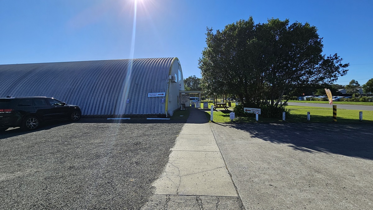 To the left is a large, dome-shaped metallic building with a corrugated surface, next to which a black SUV is parked. To the right, there is a grassy area dotted with trees and small signposts. In the foreground, a concrete path runs centrally, leading to a gravel driveway. The area around the building is covered with grass and gravel, with trees also visible.