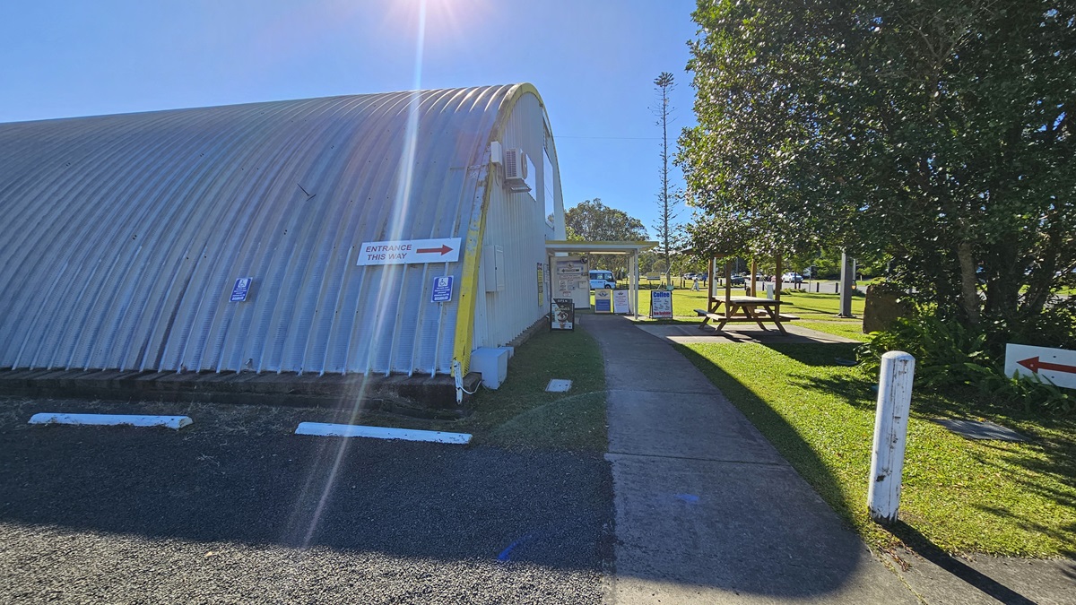 A closer view reveals a large, dome-shaped metallic building with a corrugated surface to the left. To the right, there's a grassy area with trees, small signposts, and a picnic table. In the foreground, a concrete path runs centrally, leading to a gravel driveway. The vicinity of the building is covered with grass and gravel, and trees are also visible. Directional signage is affixed to the side of the building.