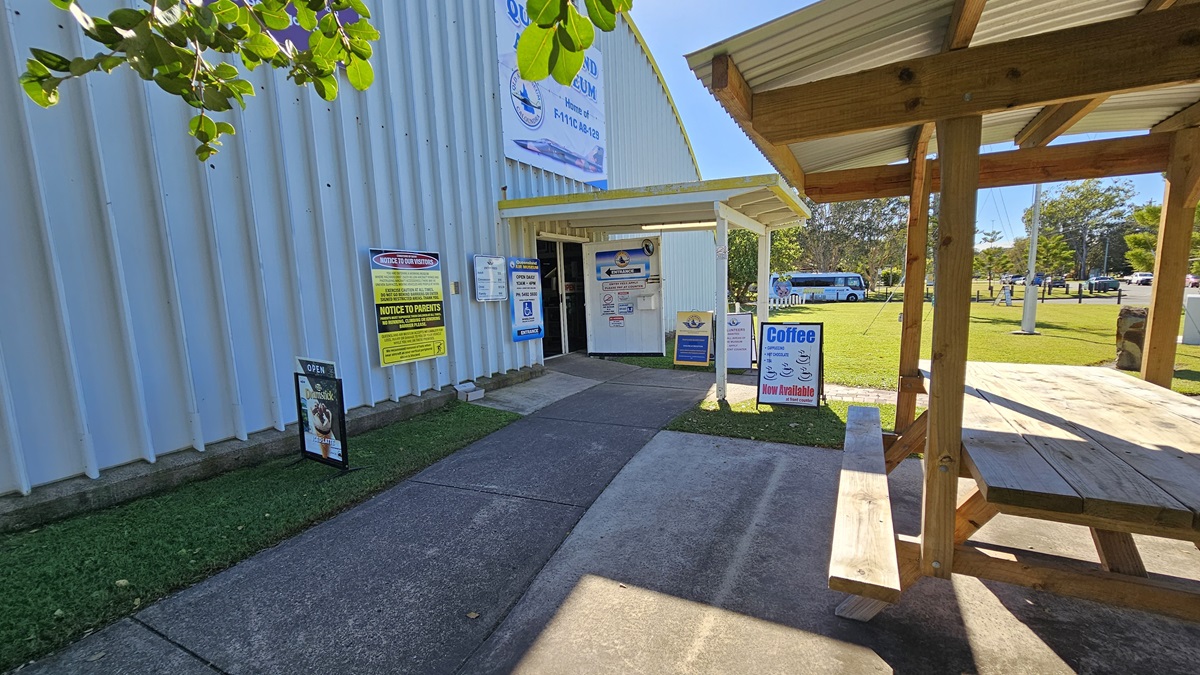 A corrugated metal building is on the left, featuring several signs near its entrance, including a "Museum Entrance" sign. On the right, there is a wooden picnic table under a metal-roofed shelter. In the background, daylight reveals a grassy area with trees and additional signage. The ground consists of a concrete path leading towards the building entrance and extending towards the picnic table.
