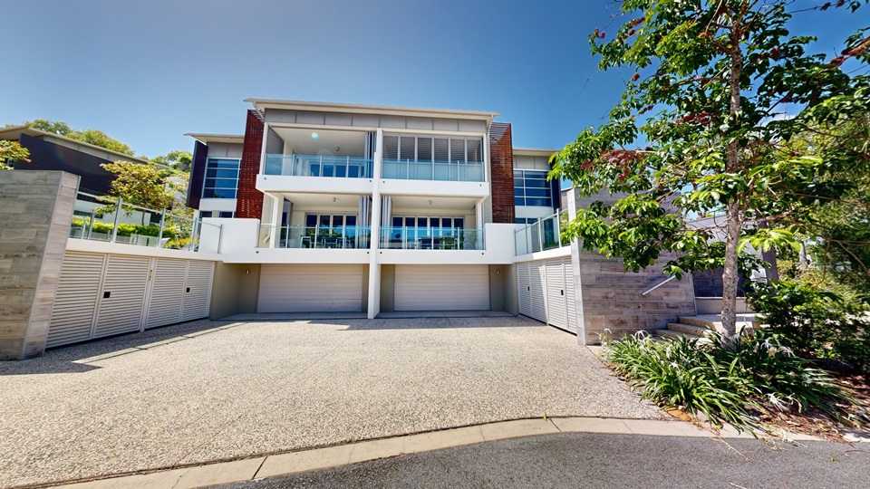 A three-story twin-style building made of concrete and glass, with a spacious front and a garage. Steps with handrails on the right lead to the upper deck. Tall trees and flowering plants adorn the front landscape.