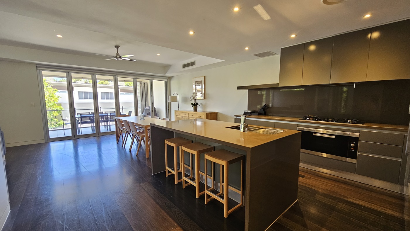 In the kitchen, a central island with a sink, is surrounded by cooking equipment and cabinets. Adjacent is a dining table with a display cabinet nearby. A framed decoration adorns the white wall. Beyond, tables and chairs are visible on the balcony. The floor is polished timber, and the wall and ceiling are white.