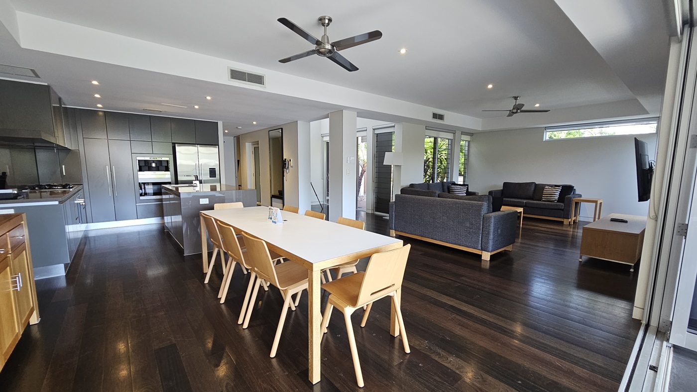 In the kitchen, a central island with a sink, is surrounded by cooking equipment, cabinets, and a fridge. Adjacent is a dining table with a display cabinet nearby. A framed decoration adorns the white wall. Beyond, tables and chairs are visible on the balcony. The floor is polished timber, and the wall and the white ceiling feature a cove design with pin lights and ceiling fans. To the right is a seating area with cushioned chairs and a large flatscreen TV.