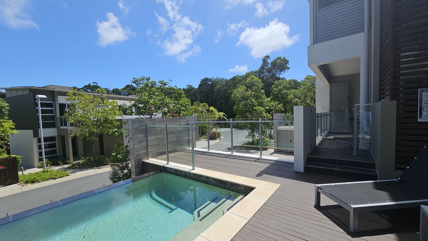 A plunge pool with steps and black sun loungers on a timber deck. Glass railing secures the area. The building facade, a blend of concrete, glass, and timber, is on the right. Trees, plants, and other villas are visible below.