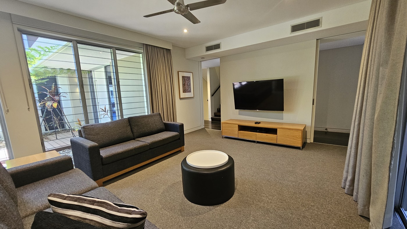 The lounge area consists of sofas arranged around a round center table facing a large flatscreen TV on a TV stand. Floor-to-ceiling grey curtains hang on both sides. A glass wall on the left provides a view of plants and trees outside. A framed decoration hangs on the left wall. Stairs with handrails lead upwards on the left. The floor has grey carpet, and the ceiling is white with a mounted ceiling fan.