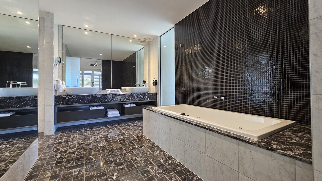 A spacious bathroom with a tub on the right. The right wall features small black tiles. The floor has printed black tiles matching the lavatory. In the middle, a large mirror sits above two hand basins with a towel organizer underneath. Liquid soap dispensers are mounted to the wall. Another large mirror is placed on the left, facing the tub. The ceiling is all white with LED lights and a ceiling fan.