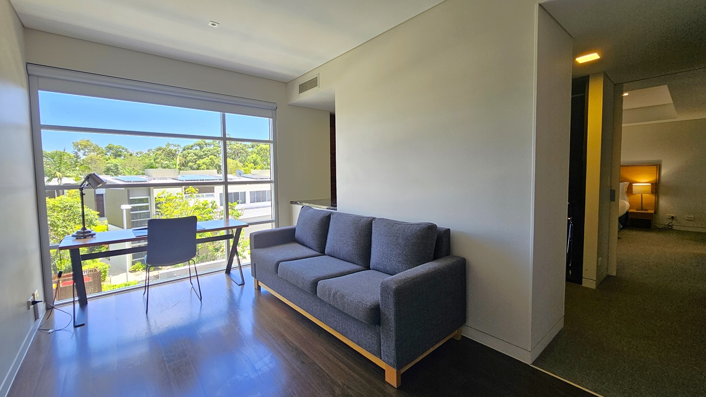An office table and chair are positioned facing the glass wall, offering a view of the villas and trees. A desk lamp is on top. To the right is a grey sofa. The floor is polished timber. Adjoining the room is the bedroom with grey carpets, white walls, and a white ceiling with LED lights.