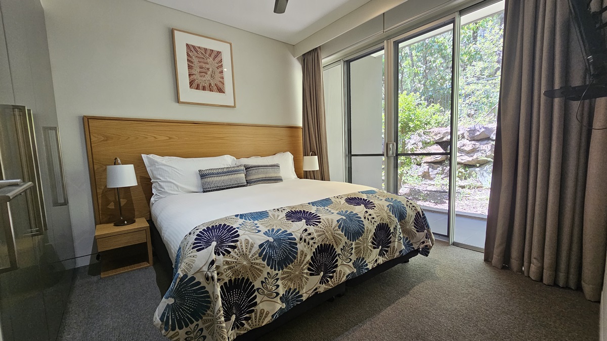 On the right is a king-size bed with printed linens and a wooden headboard, flanked by matching bedside tables and lampshades. To the right is a glass partition, with floor-to-ceiling grey curtains drawn to both sides. The floor is carpeted in grey, and the walls are white with a photo frame. A flat-screen TV is mounted on the wall. To the left is a glass door.