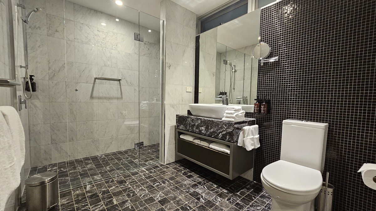 To the right is a toilet bowl positioned against the tiled wall. A tissue dispenser and a liquid soap dispenser are on both sides. To the left is a glass door securing the shower area. Shower and hand grabs are mounted to the wall, along with toiletries. A rubbish bin and a white towel are placed nearby. Adjacent is a lavatory with a hand basin and folded white towels on top. A towel organizer is placed underneath. The floor has printed black tiles, and the ceiling is white.