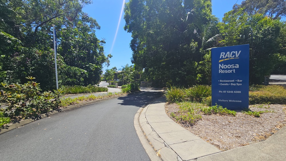 An asphalt road flanked by trees, shrubs, and grass. A concrete pathway veers to the right. In the middle is a blue sign displaying the facility name atop a concrete planter. A tall street light stands on the left.