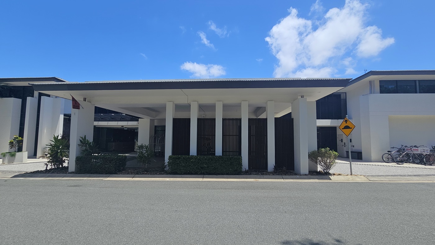 A single-storey building with a white corrugated roof, bordered by a black metal fence. Two-storey buildings flank both sides. Bicycles for hire are parked to the right against the building. Neatly trimmed shrubs line the front. A yellow street sign stands to the right.