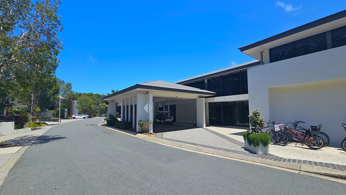 A contemporary building of concrete and glass. Luggage trolleys and carts are near the driveway. Neatly trimmed shrubs and plants line the front. Bicycles for hire stand against the white building on the right. Tall trees border the asphalt road on the left. Further ahead, two white cars parked in front of the buildings are visible.