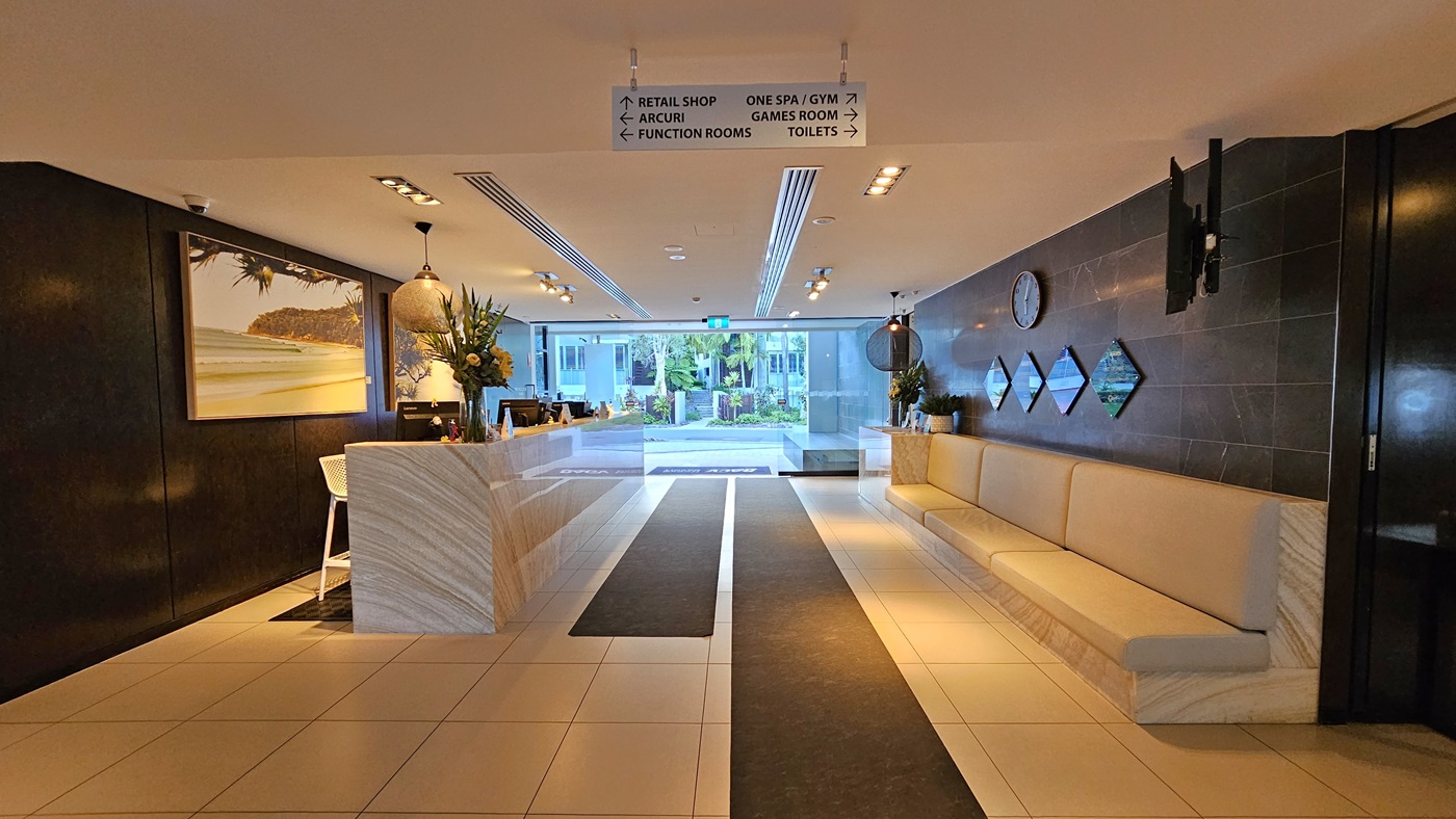 A view back towards the entrance doors with the reception on the left, two brown runners in the middle and a cream couch lining the wall to the right. Awards and presentations hang on the wall on the right.