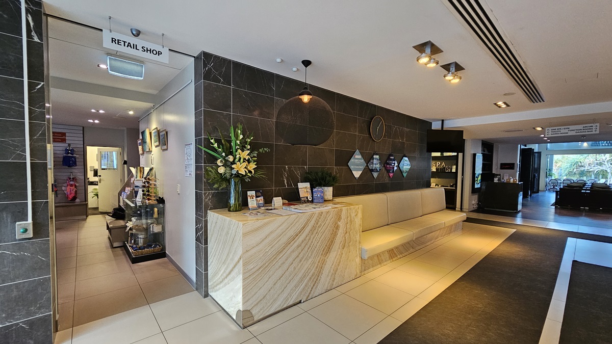 The entrance to the retail shop to the left. On the right of this entrance is a cream couch lining the wall, A table with flowers and brochures. The wall features awards and presentations.