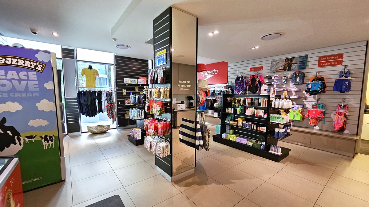 The retail shop offers a variety of swim gear for all ages, along with toiletries and bags on display shelves and walls. T-shirts are showcased on the glass wall. An ice cream freezer is on the left, beside a wall with an advertising sign. The ceiling and floor are white with LED lights.
