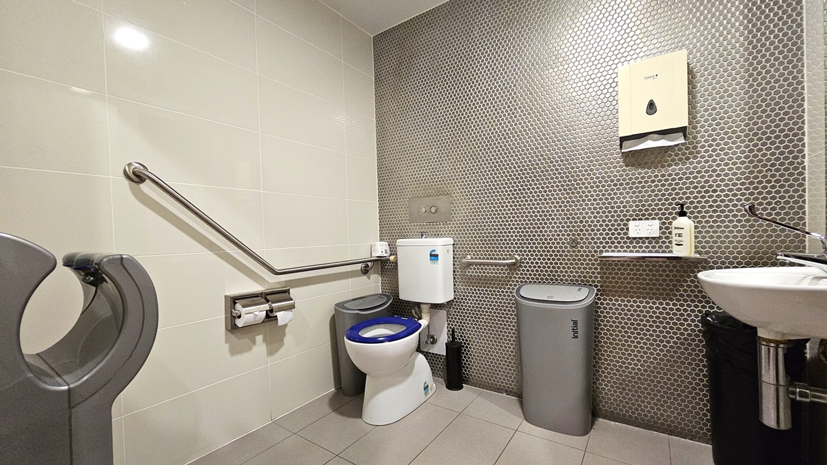 The accessible toilet features a toilet bowl with a hand rail, located in the left corner. A tissue dispenser and a rubbish bin are placed beside it. To the right, a hand basin is mounted against the wall, with a rubbish bin underneath. Adjacent to it, a ledge holds liquid soap and a tissue dispenser. The wall is adorned with white and grey tiles with a honeycomb design, while the floor is tiled in cream.