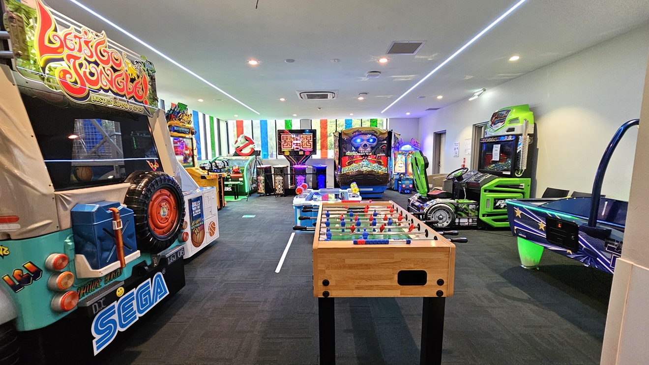 In the middle are electronic arcade games and driving machines, with hockey machines and football tables on the right. A large riding machine is also on the right. The floor is carpeted with tiles, and the walls feature colourful linear accents, while the ceiling is white with long fluorescent lights.