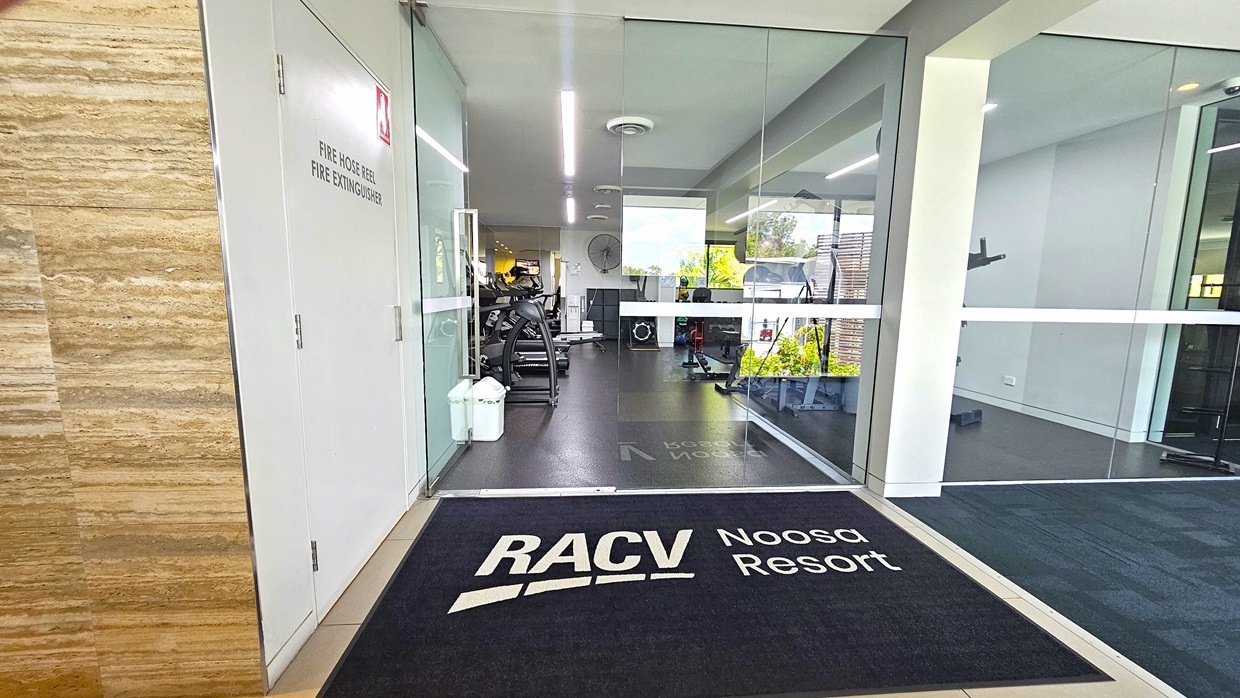 In the centre, there's a manual outward-opening glass door with a black mat displaying the facility's name. On the left wall, there's a fire hose reel and a fire extinguisher. The exterior wall has a timber design. Inside, the gym floor is covered with black rubber tiles. Gym equipment is arranged in the middle, and a white rubbish bin is placed near the entrance.