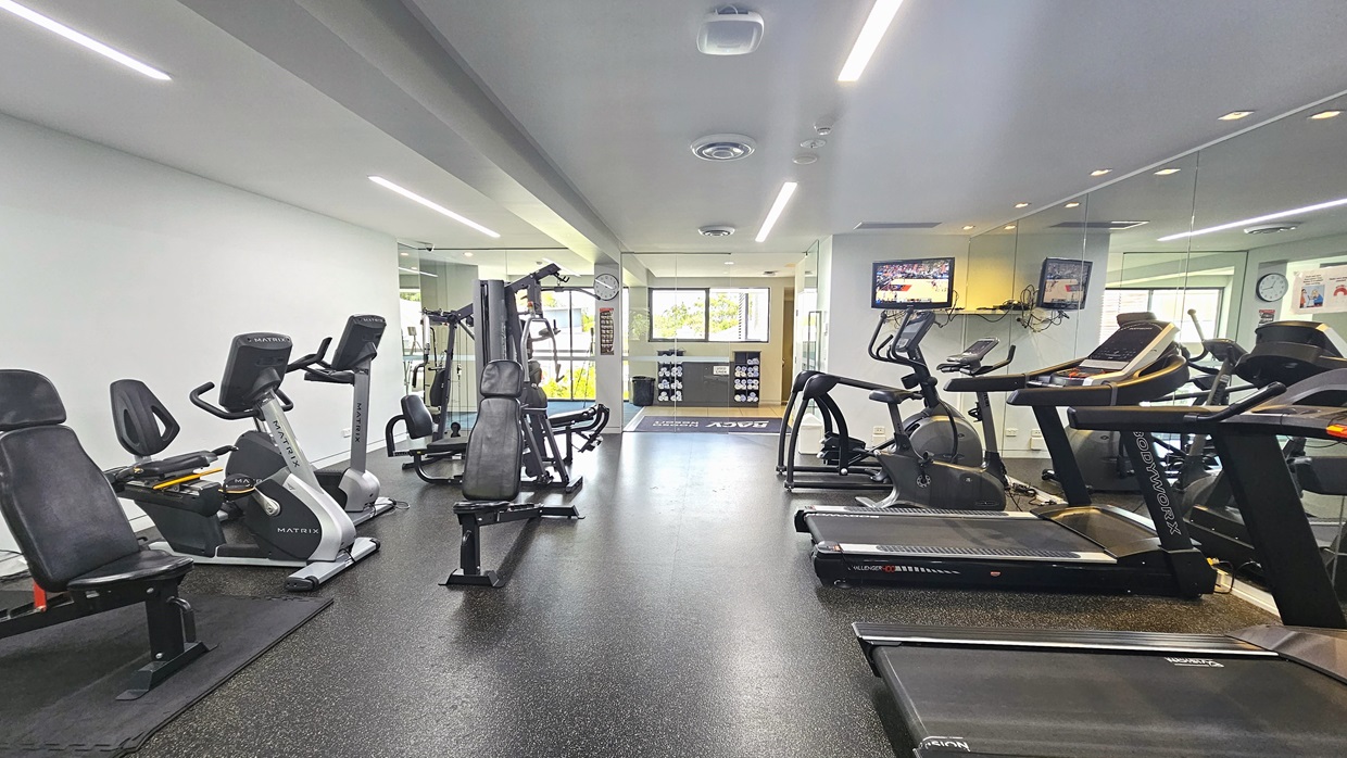 To the left, lat pulldown machines, bench chairs, and other machines are arranged. To the right, rows of treadmills and step machines. Flatscreen TVs and a wall clock are mounted on the middle wall. The floor has black rubber tiles, with white walls and ceilings featuring fluorescent lights. At the far end is the entrance door. Rolled white towels on a display rack are visible.