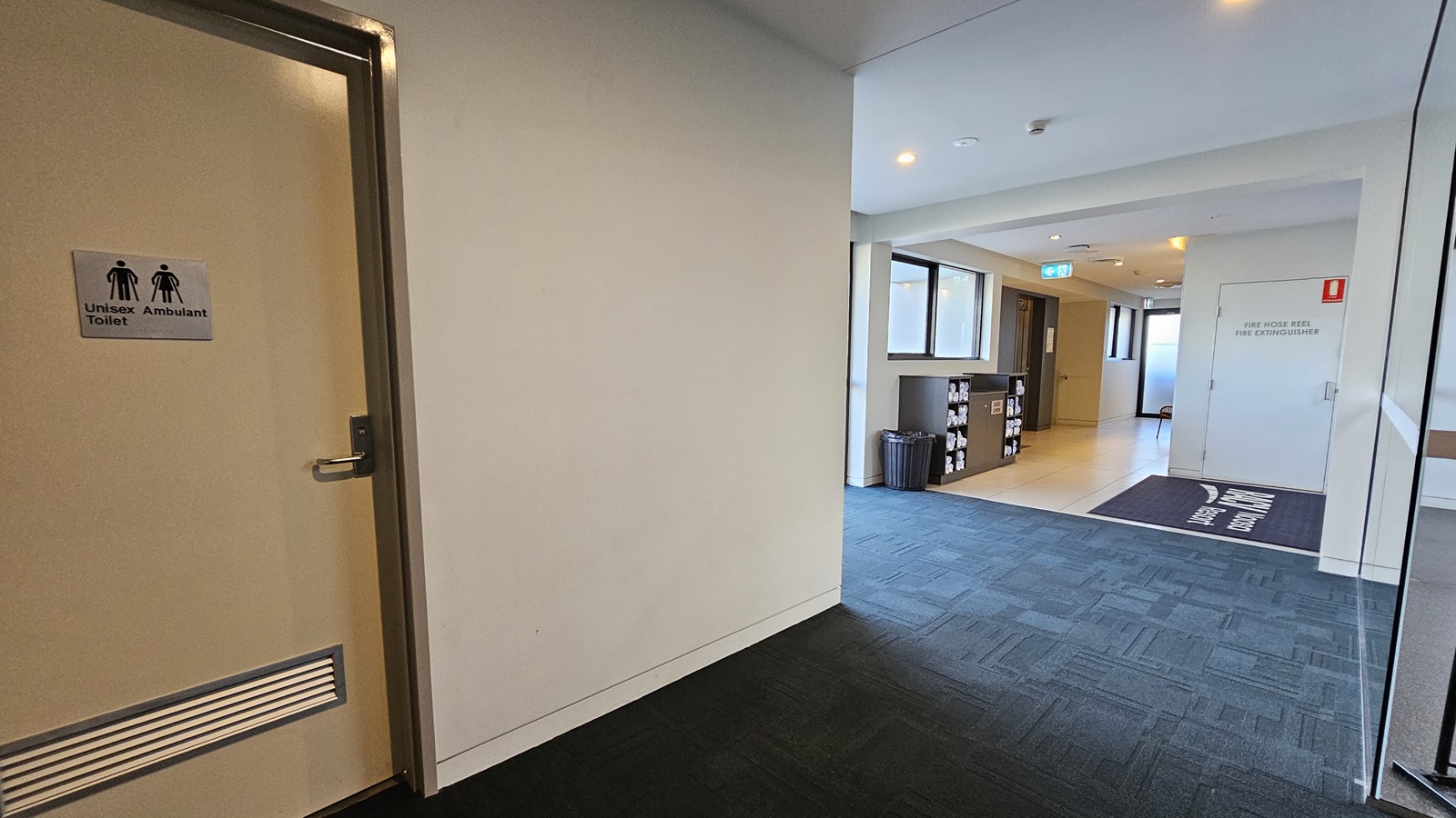 To the left is a Unisex Ambulant Toilet. To the right is a glass door that opens outward. A black matting bearing the facility's name is positioned on the door near the entrance. Further to the left is a display rack with rolled white towels against a glass window. A black rubbish bin is beside it. The floor transitions from grey to black rubber tiles inside.