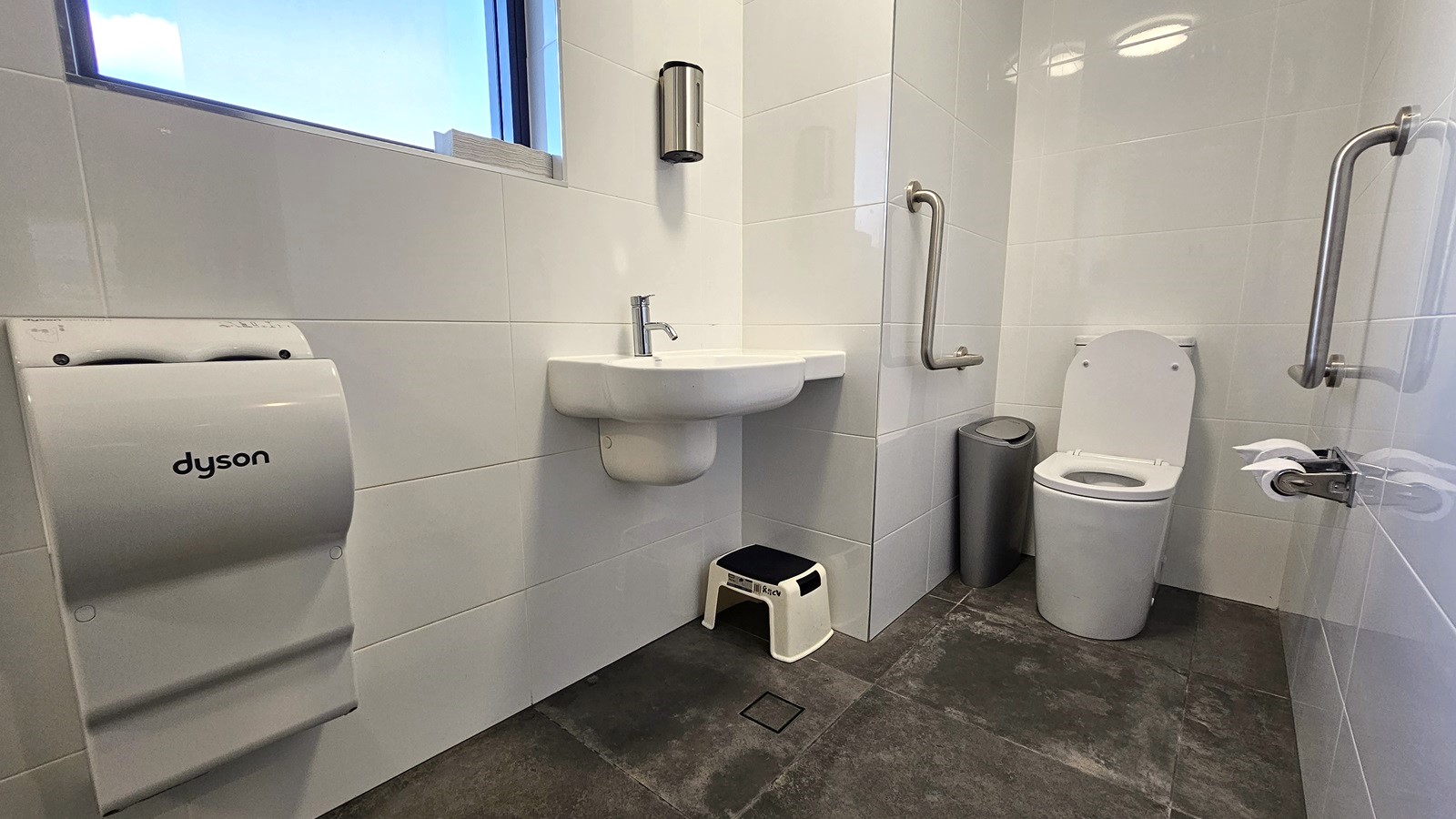 Inside the Ambulant Toilet, a toilet bowl with handrails is in the corner. On both sides are tissue holders and rubbish bins. A hand basin is mounted on the left wall with a liquid soap dispenser above and a small plastic stool underneath. Next to it is a hand dryer mounted on the wall near the glass window. The wall has white tiles, and the floor has grey tiles.