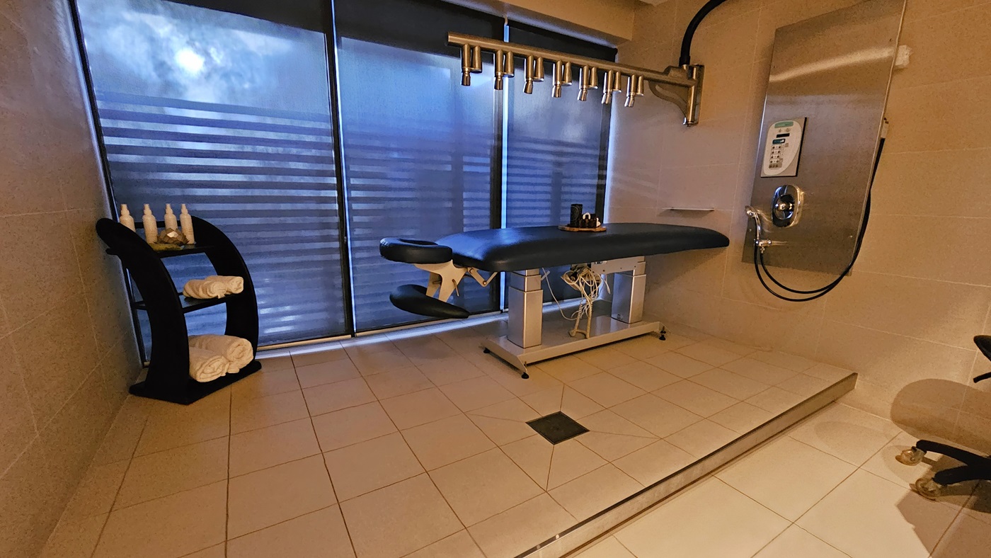 On the left corner, a black shelf holds rolled white towels. Next to it, is a table with an adjustable headrest. Light fixtures are on the right wall, beside a shower. Glass partitions in the middle and on both walls have brown tiles matching the floor.