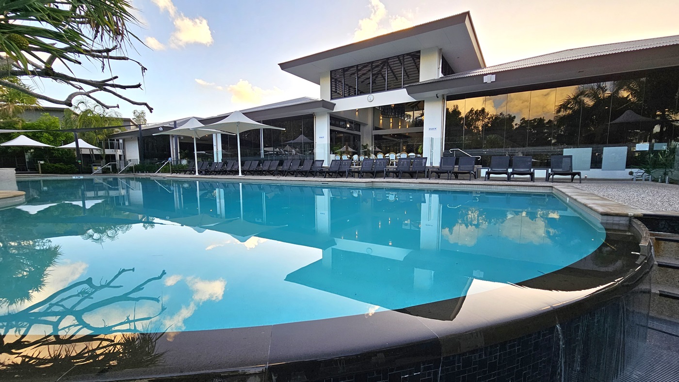 In the middle is a large pool with sun loungers lining the pool deck. A building made of concrete and glass stands behind it. Large white umbrellas cover a section of the pool. To the right is a staircase that leads to the upper section of the pool deck. A tree casts its shadow on the pool water.