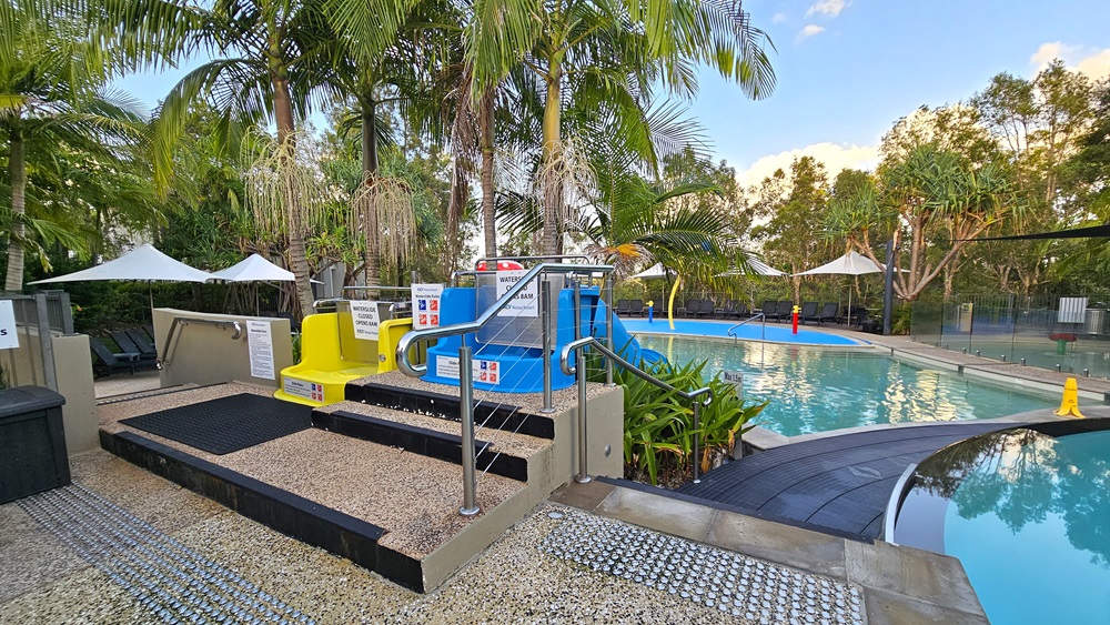 On the right are two pools separated by a ramp with handrails and truncated domes. On the left, a stepped entry leads to blue slides connected to the kids' pool. Large white umbrellas shade sun loungers on the pool deck. Palm trees encircle the area.