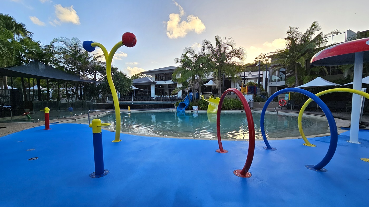 In the middle is a children's pool with steps and handrails. Behind it is a yellow and blue slide. In front is a splash area with a vibrant blue non-slip floor. Mushroom umbrella showers, multicolored sprinklers, and hoops are positioned. Shade cloth covers the left section of the pool. Tall palm trees surround the pool area. Further behind is the facility building.