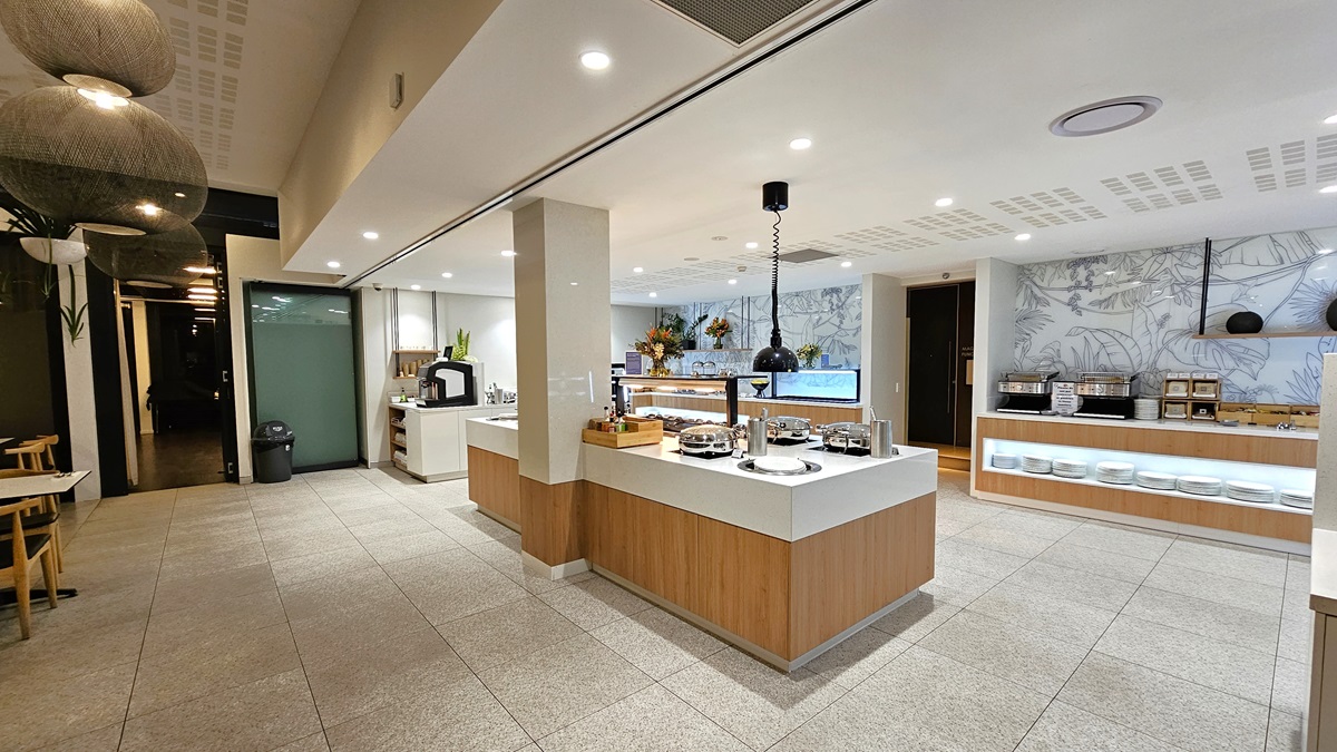 In the middle, a buffet section displays food items. Adjacent, another counter showcases silverware on shelves and a food display rack on top. Movable chairs and tables are to the left. A door on the left leads to another buffet section. The white ceiling features stylish light fixtures and hanging plants. The right wall has an intricate design. The floor has cream tiles matching the wall.