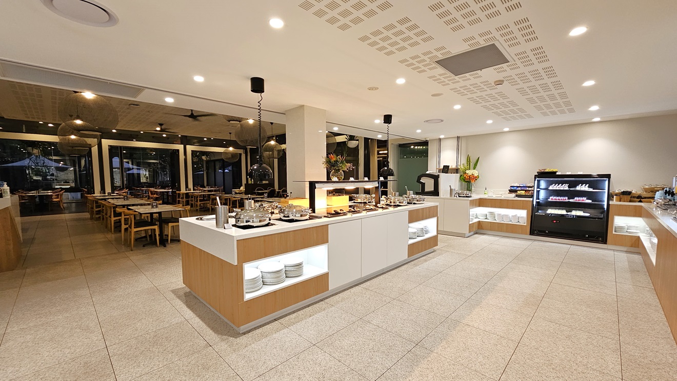 In the middle, a buffet section displays food items. Adjacent, another counter showcases silverware on shelves and a food display rack on top. A fridge displaying food items is placed in the middle. Movable chairs and tables are arranged to the left. The white ceiling features stylish light fixtures and ceiling fans. The floor has dotted cream tiles matching the wall. A glass wall borders the left section.
