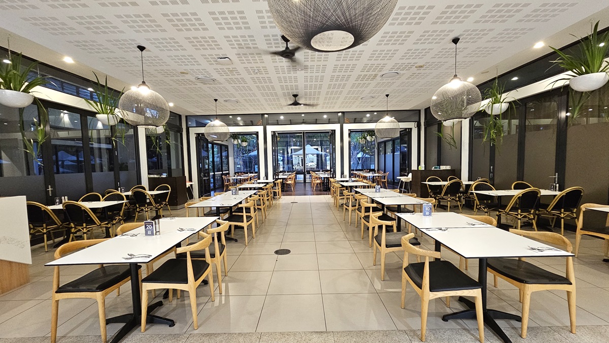 Movable chairs and tables are arranged in rows in the middle and on the sides, with utensils laid out. The white ceiling has a slit design with ceiling fans and stylish light fixtures resembling giant balls of thread hanging down. Ornamental plants also hang from the ceiling on both sides. The floor is tiled with white, and the area is surrounded by glass walls.