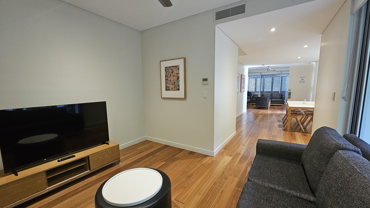 On the left, a flatscreen TV sits atop a TV stand. To the right, grey cushioned chairs are arranged against a glass wall. Further to the right, a dining table with wooden chairs and another lounge with grey cushioned chairs. The ceiling is all white with LED lights and a ceiling fan. The white wall features a framed decoration. The floor has timber designs.