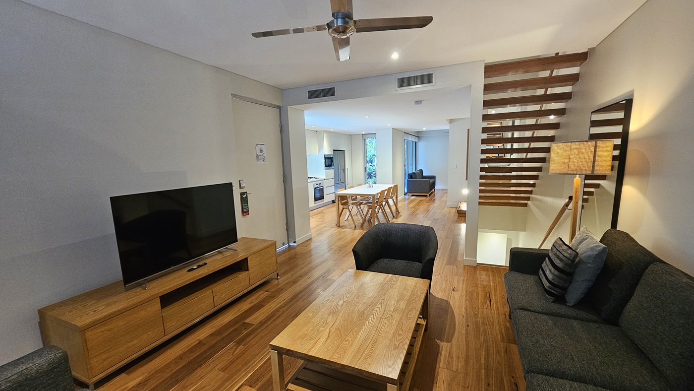 To the left, a flatscreen TV sits atop a TV stand. Grey cushioned chairs are arranged against the wall on the right. A wooden table is placed in the middle. Further to the right, is a dining table with wooden chairs and another lounge with grey cushioned chairs. A wooden step is also positioned on the right, underneath is a lampshade and a mirror. The ceiling is all white with LED lights and a ceiling fan. The floor has timber designs.