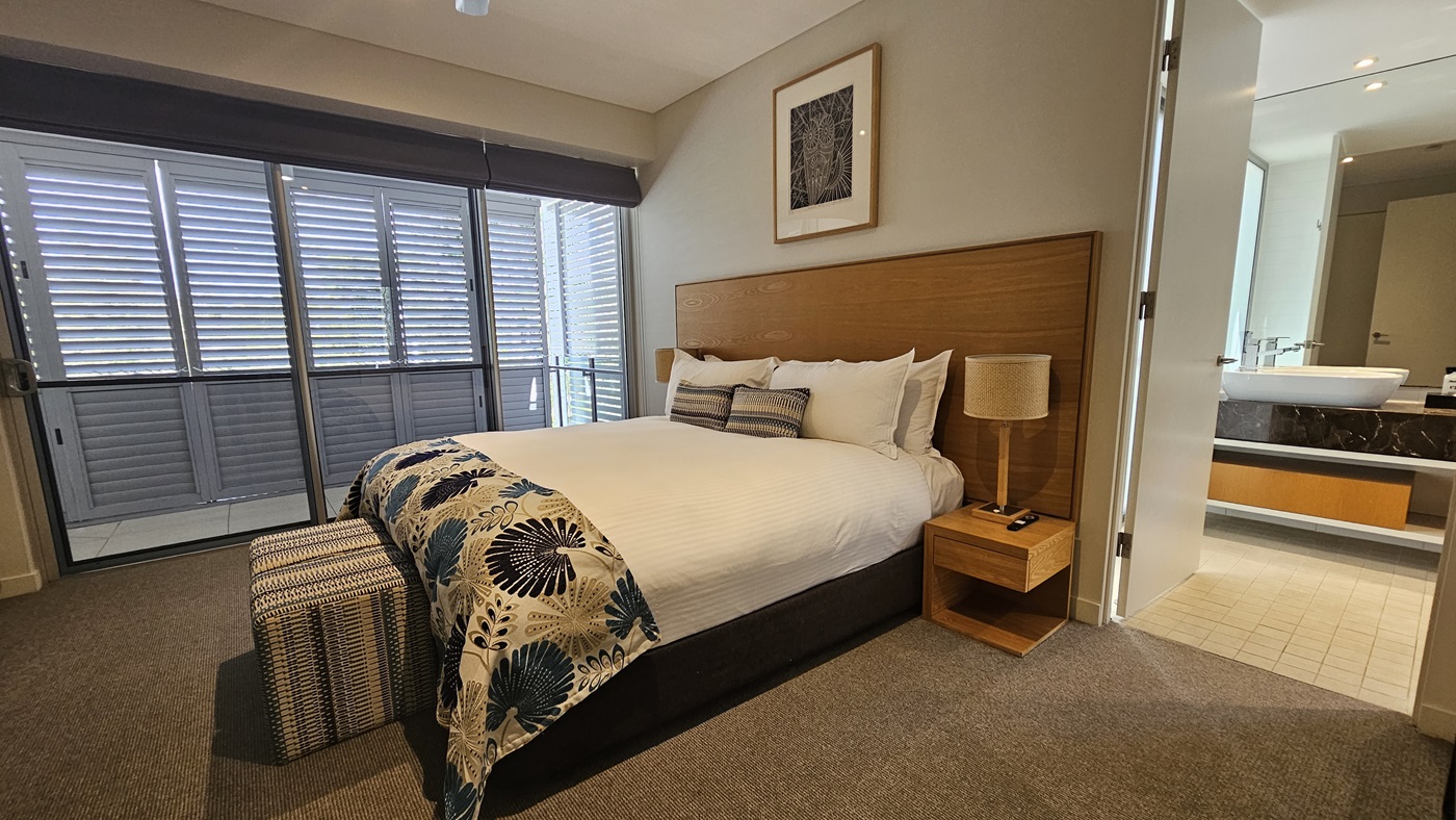 To the right, a king-size bed with a wooden headboard and matching bedside tables. Lampshades top the tables. The toilet, equipped with a hand basin and a large mirror is visible to its right. To the left, retractable curtains on the left wall. A framed decoration hangs on top of the bed. The floor has grey carpet and the ceiling and walls are white.