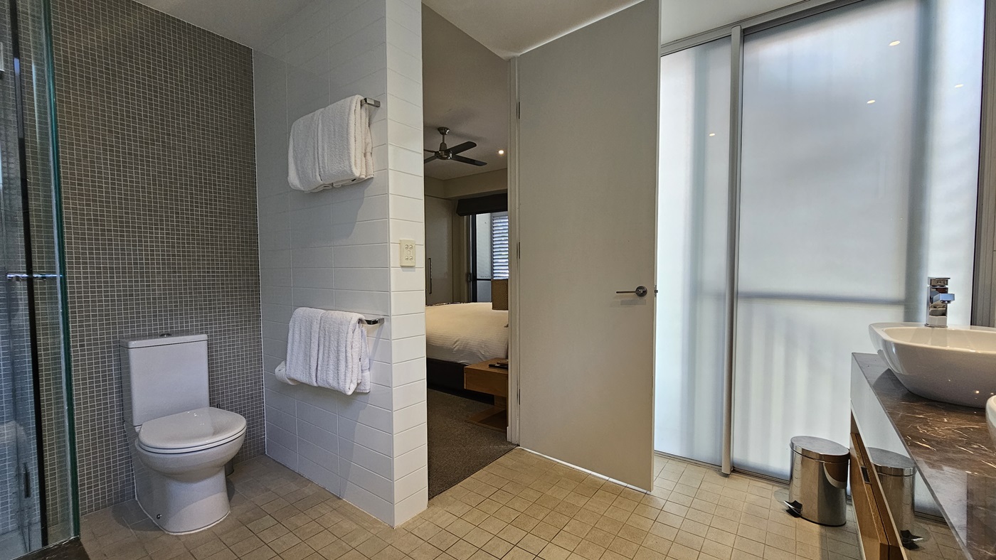 To the left, a toilet bowl is positioned against the grey wall. Towels hang on a handrail to the right. A glass wall is to its left, while a hand basin stands to the right, in front of a mirror. A rubbish bin is nearby. The wall on the right is made of glass and white and grey tiles in the middle. The floor has cream tiles. A section of the bedroom is visible.